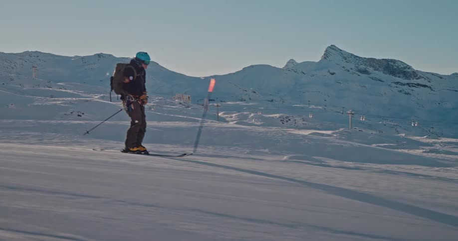 A professional skier with a backpack carves down a wide, sun-drenched mountain slope in epic slow motion