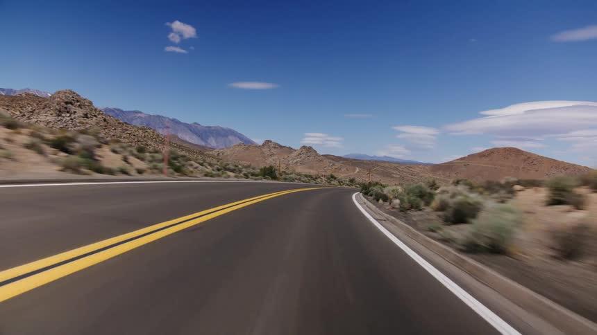 A scenic point-of-view drive on a winding desert road towards snow-capped mountains under a clear blue sky