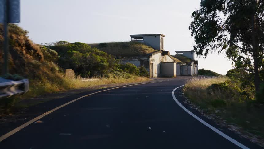 A smooth tracking shot moves along a winding coastal road, revealing an old, abandoned military bunker built into the hillside at sunrise