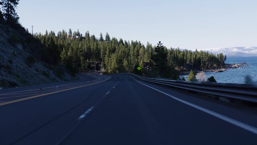 A first-person point-of-view driving along a scenic highway next to a beautiful blue lake with a pine tree forest on a sunny day