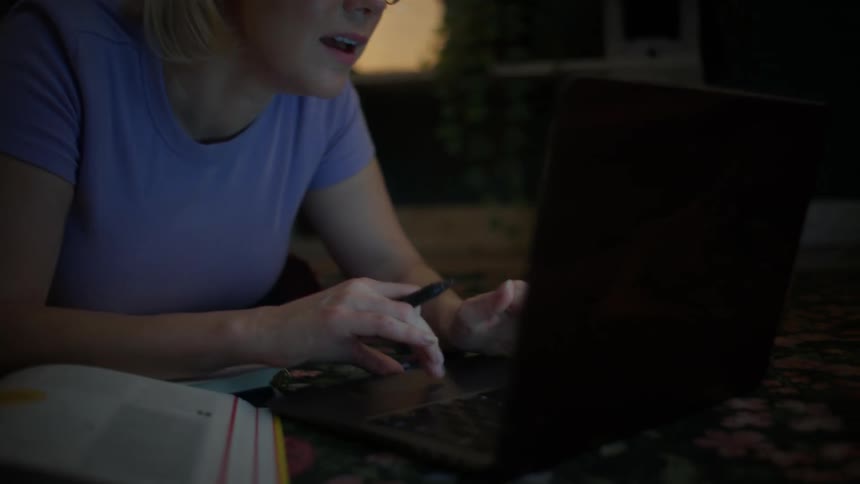 A focused young woman wearing glasses scrolls intently on her laptop while studying late at night in a dimly lit room