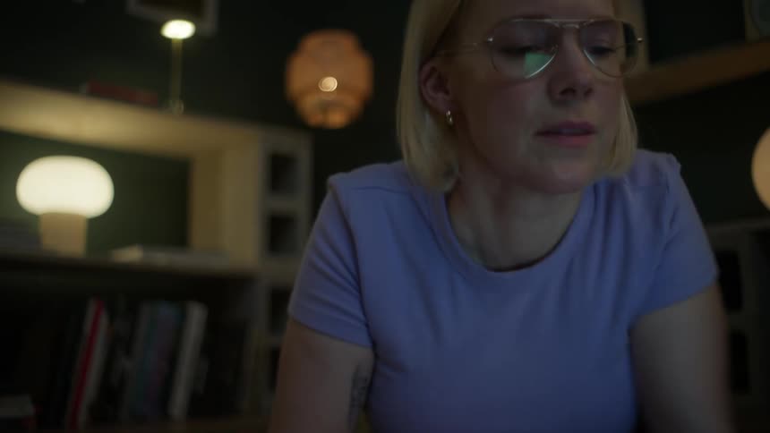 A young blonde woman with glasses studies from a large book while lying on the floor of her cozy, dimly lit apartment at night