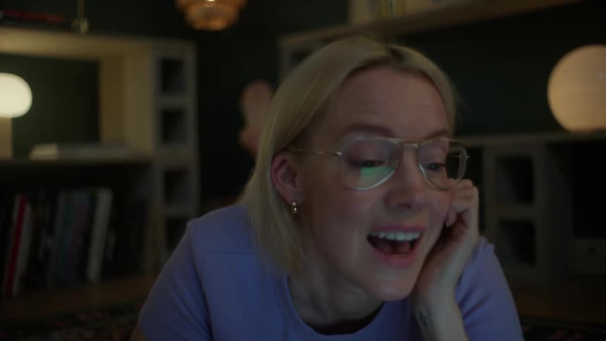 A happy blonde student with glasses enjoys a video call on her laptop while relaxing on the floor in a cozy, dimly lit room