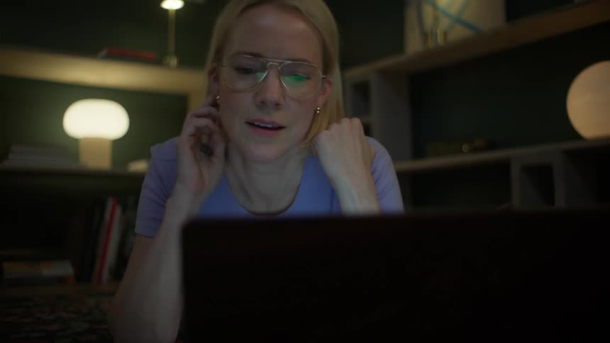 A focused blonde woman in glasses has a late-night video call on her laptop while lying on the floor in her cozy living room