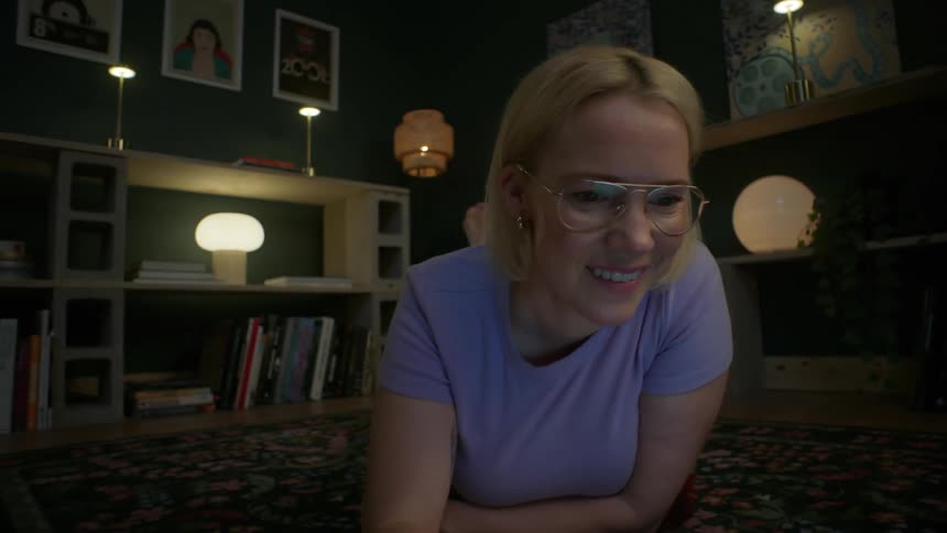 A slow pull-back shot reveals a smiling young woman with glasses using her laptop while comfortably lying on the floor in a cozy, modern apartment at night
