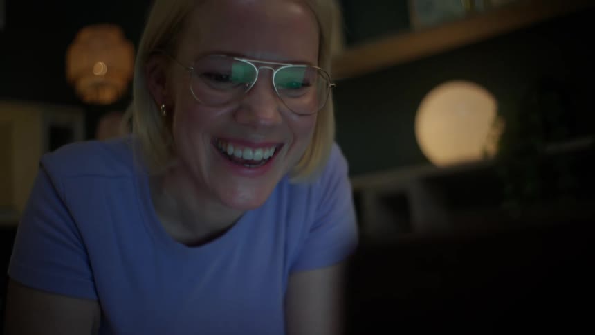 A slow tilt-up reveals a happy young woman with glasses laughing while using her laptop late at night in a cozy room
