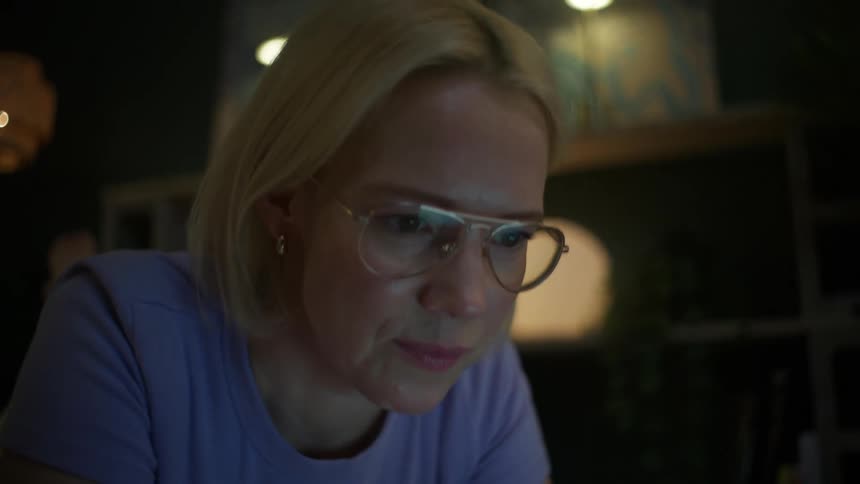 A woman with glasses works on her laptop and takes notes while lying on the floor in a cozy, dimly lit room at night