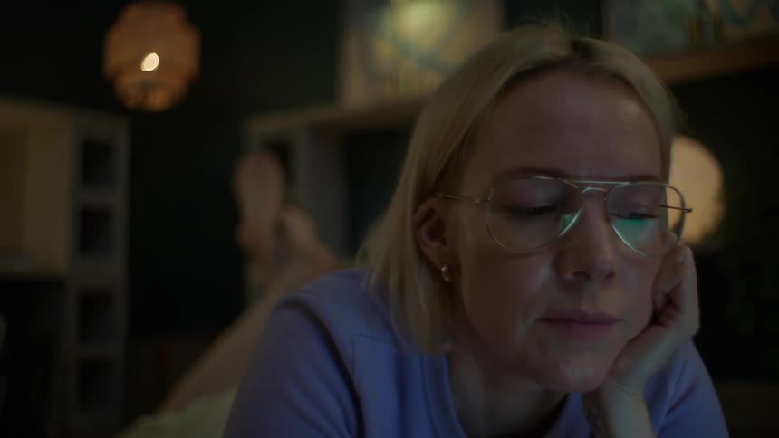 A young blonde woman wearing glasses studies late into the night, lying on the floor of her cozy apartment with her laptop and textbooks