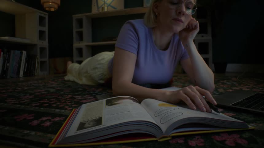 A focused woman studies on the floor of her cozy living room at night, using her laptop and a book for research