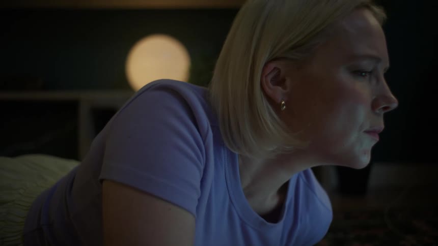 A serious blonde woman lies in a dark room at night, her face illuminated by the glow of a screen she is intently watching
