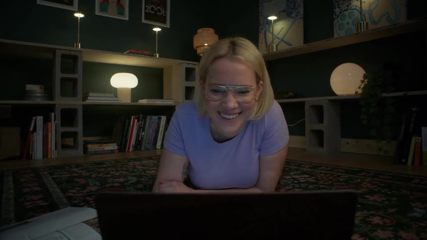 Dolly zoom on a smiling young woman watching her laptop at night while lying on the floor of her cozy living room