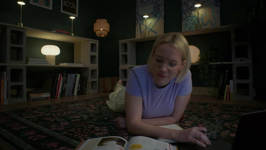 A focused young woman lies on the floor of her cozy, stylish room at night, studying with her laptop and books