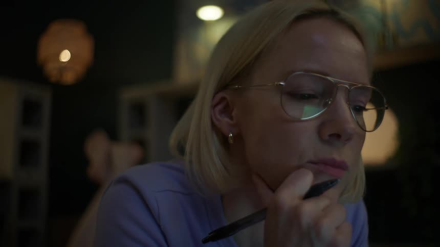 A thoughtful woman wearing glasses works on her laptop late at night, lying on the floor in a cozy, dimly lit room