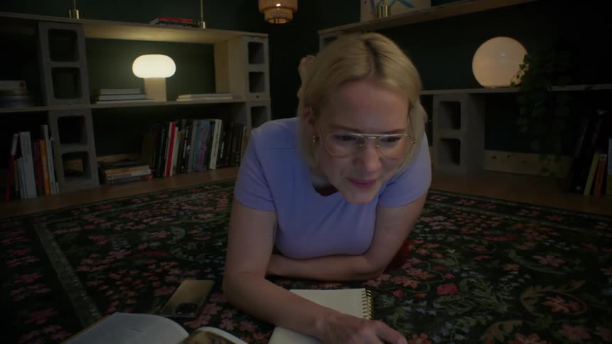 Slow dolly push-in on a smiling young woman with glasses studying on her laptop while lying on the floor at night