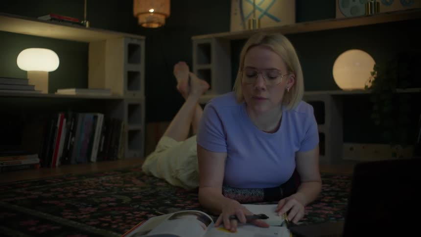 A young woman wearing glasses comfortably lies on her apartment floor at night, taking notes and using her laptop to study