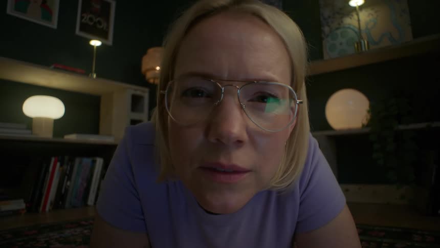 A woman with glasses lies on the floor of her dimly lit living room, her face illuminated by a screen as she watches intently and then smiles