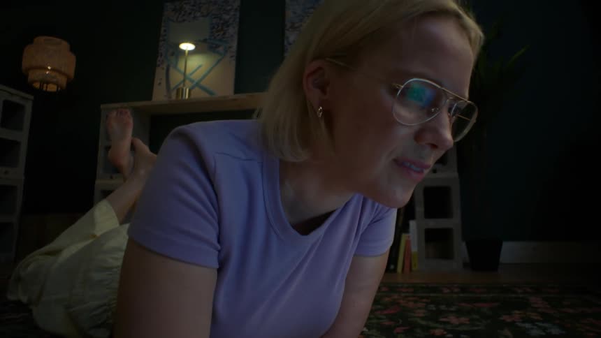 A slow dolly shot captures a young woman with glasses smiling while studying on her laptop, lying on the floor at home in a dimly lit room