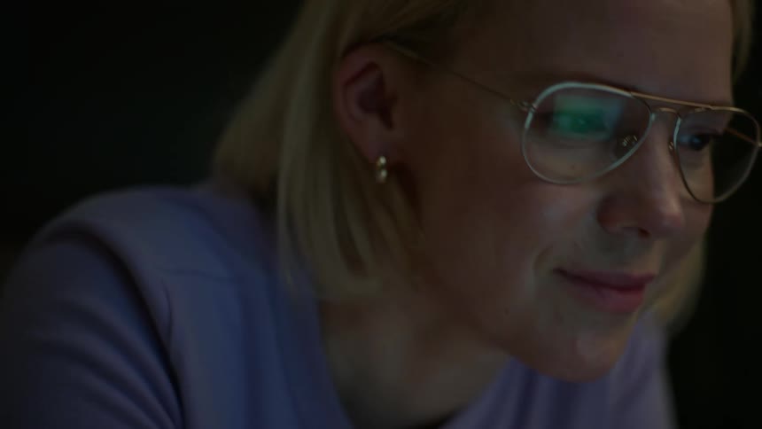 Close-up of a blonde woman wearing glasses, smiling as she works on her laptop late at night in a dimly lit room