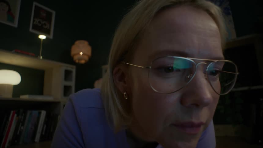 Close-up slow push-in shot of a focused woman with glasses watching a screen at night in her living room