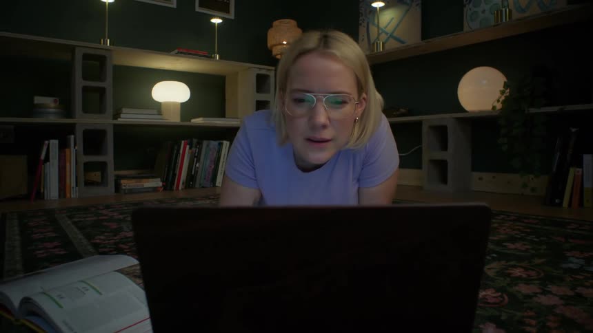 A young woman with glasses lies on the floor, intently focused on her laptop screen in a dimly lit room as the camera slowly pushes in