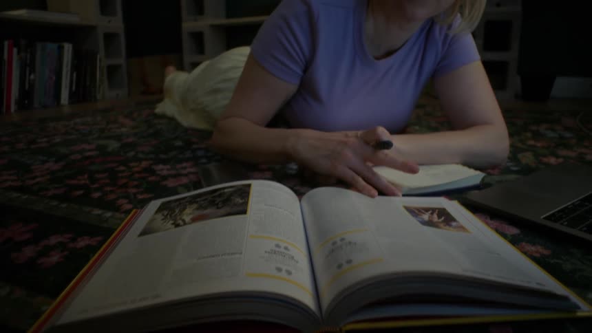 A young woman with glasses lies on a floral rug, flipping through a large book while studying at home in a dimly lit room