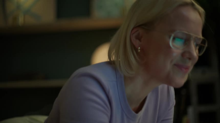 A slow focus pull reveals a smiling woman with glasses studying a book on the floor while talking on the phone