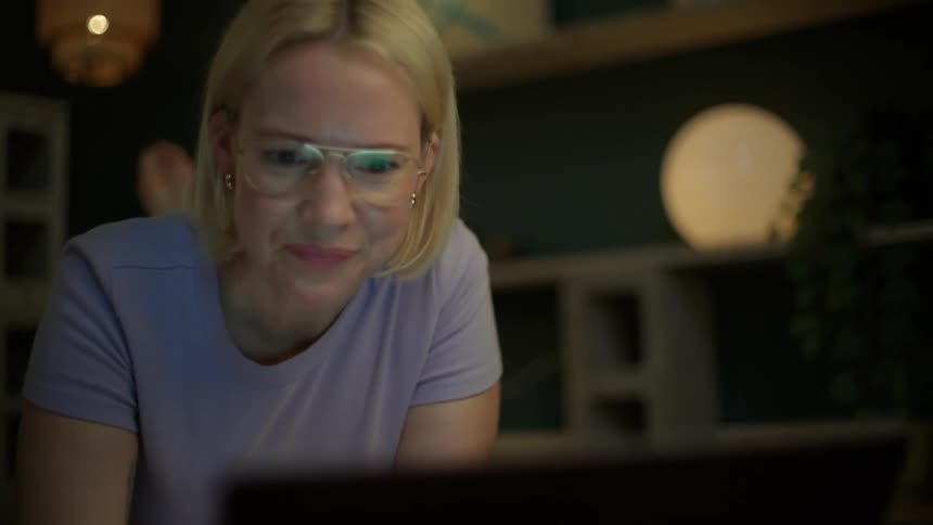 A close-up of a blonde woman wearing glasses, smiling and showing emotion while watching her laptop in a dimly lit room at night