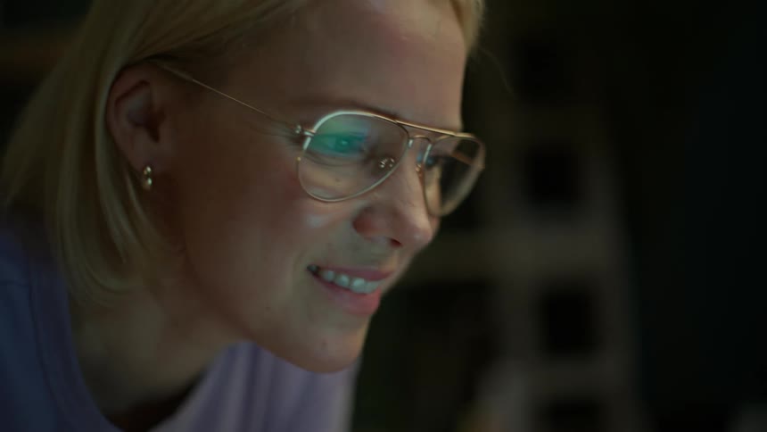 A close-up shot of a blonde woman with glasses smiling as she types and looks at her laptop screen in a dimly lit room