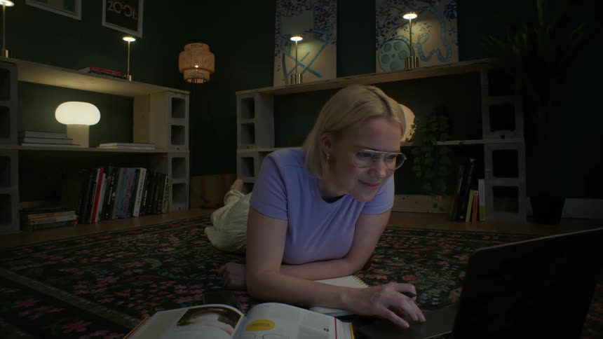 A smiling blonde woman with glasses lies on a rug in her stylish, dimly lit apartment at night while working on her laptop