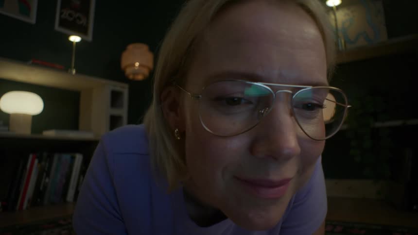 A low-angle close-up of a woman in glasses smiling as she looks at a glowing screen while lying on the floor at night