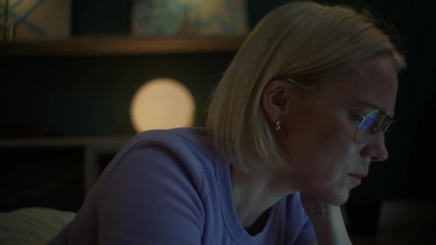 A focused blonde woman works late into the night on her laptop, eventually taking off her glasses from fatigue