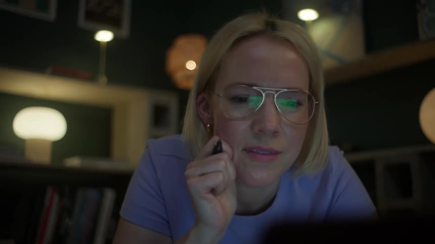 A focused young woman wearing glasses works late at night on her laptop, her expression shifting from deep concentration to a joyous smile of success