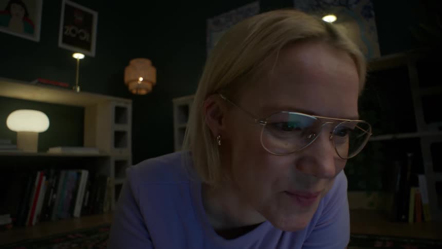 A smiling young woman wearing glasses lies on the floor of her cozy, dimly lit room at night, using her laptop computer as the camera slowly pushes in