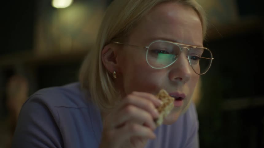 A focused woman with glasses snacks while working on her laptop at night, her serious expression turning into a smile