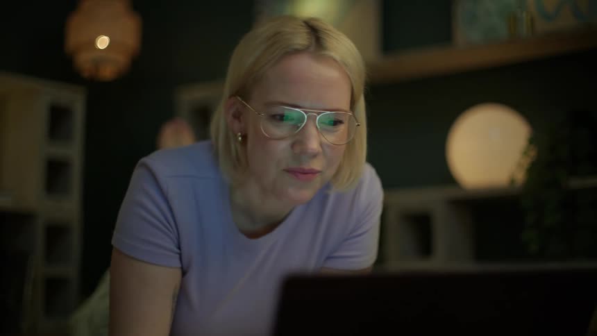 A thoughtful woman with glasses scratches her head while focusing on her laptop screen in a dimly lit room at night