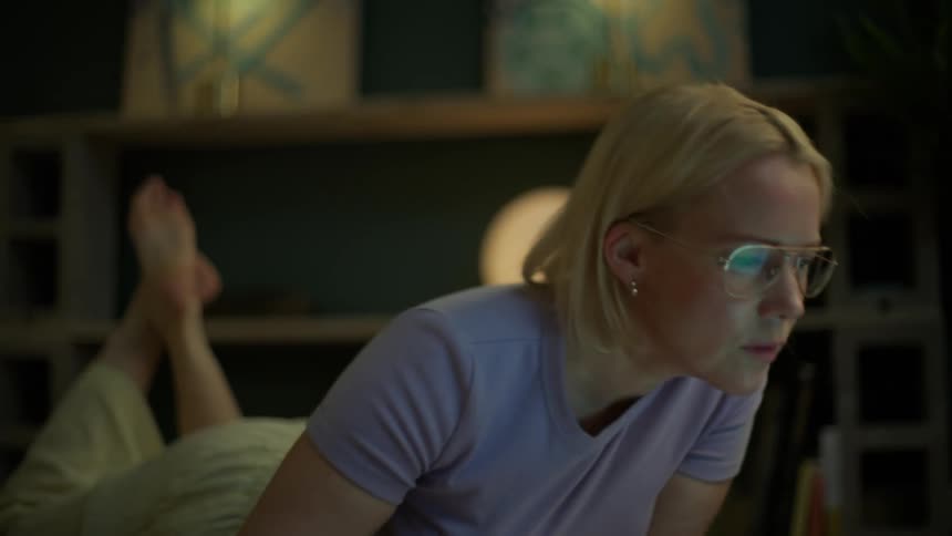 Young blonde woman with glasses studies on the floor with a book and notebook before happily working on her laptop at night
