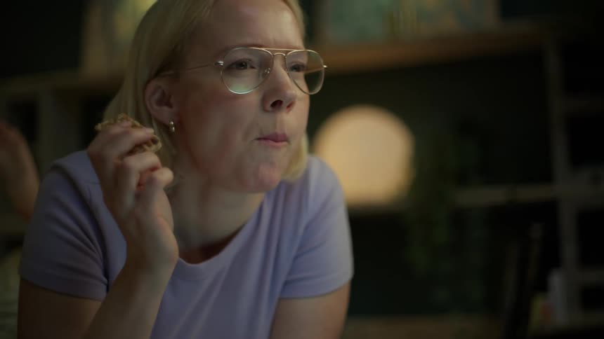 A focused blonde woman in glasses lies on her bed, eating a granola bar while working or studying on her laptop late at night