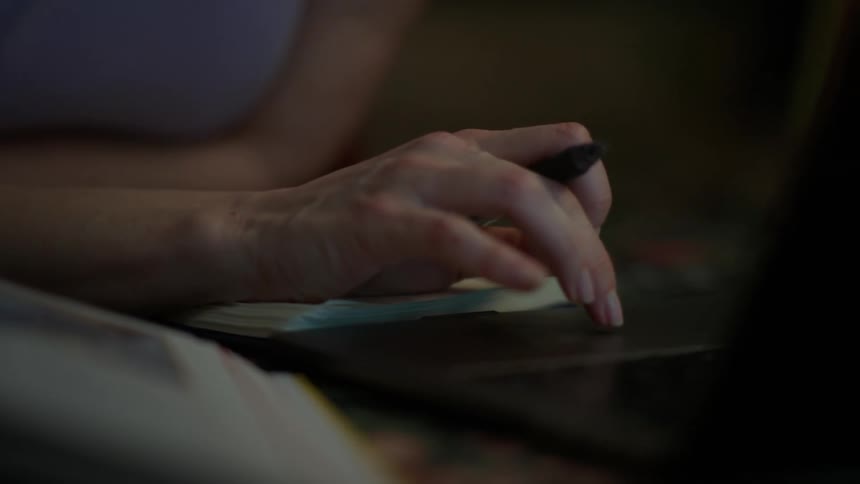 A focused young woman with glasses works on her laptop late at night, her face illuminated by the screen