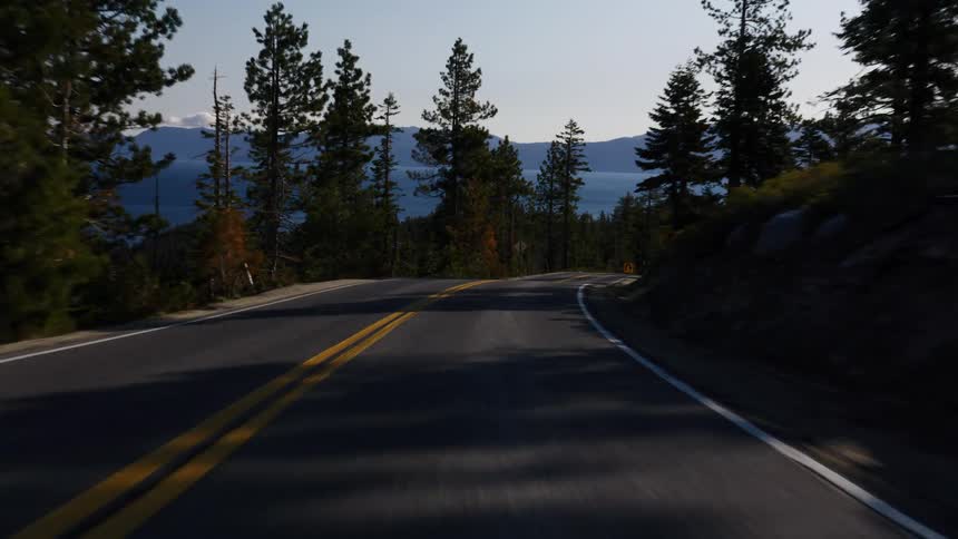 A point-of-view drive along a winding, scenic mountain road overlooking a vast blue lake through a sunlit pine forest