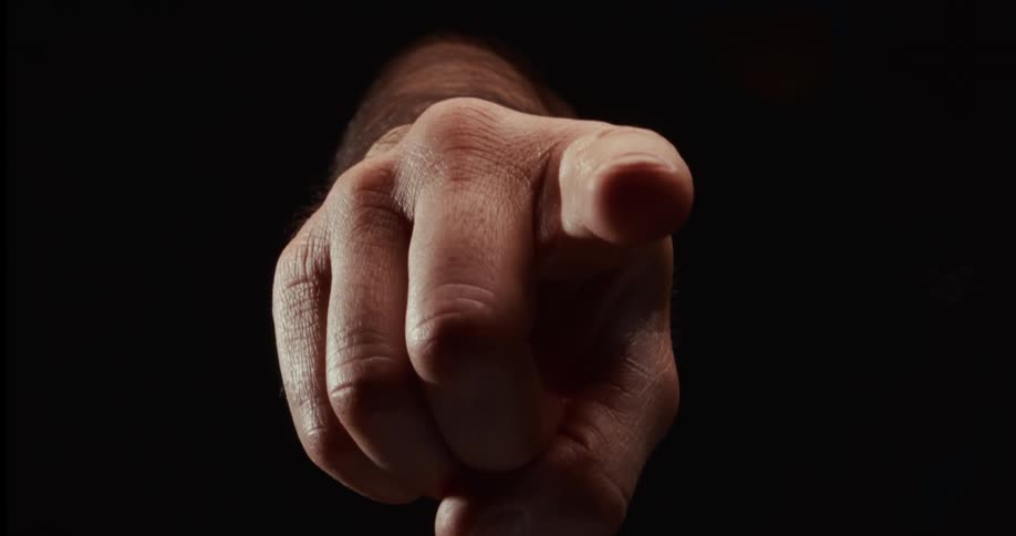 A dramatic close-up of a man's hand emerging from the darkness to point its finger directly at the camera in slow motion