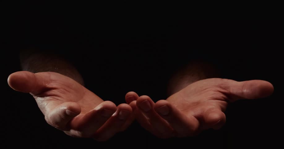 A close-up of a man's hands slowly emerging from darkness, opening in a cupped gesture of praying or giving against a black background