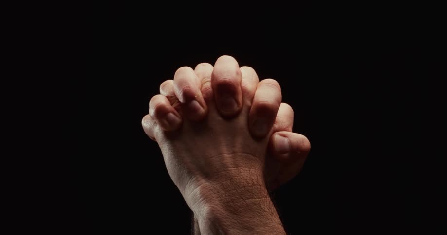 Dramatic close-up of a man's hands clasped tightly together in prayer against a solid black background