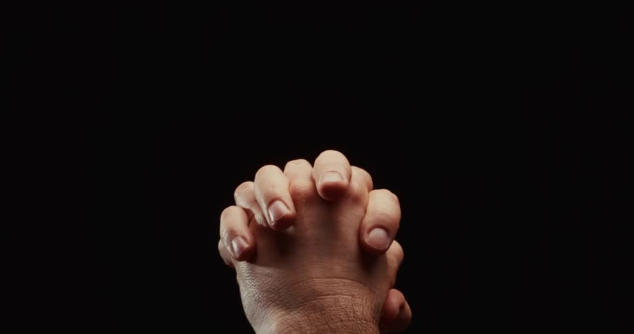 Dramatic close-up of two hands slowly interlocking fingers and clasping together in prayer against a black background