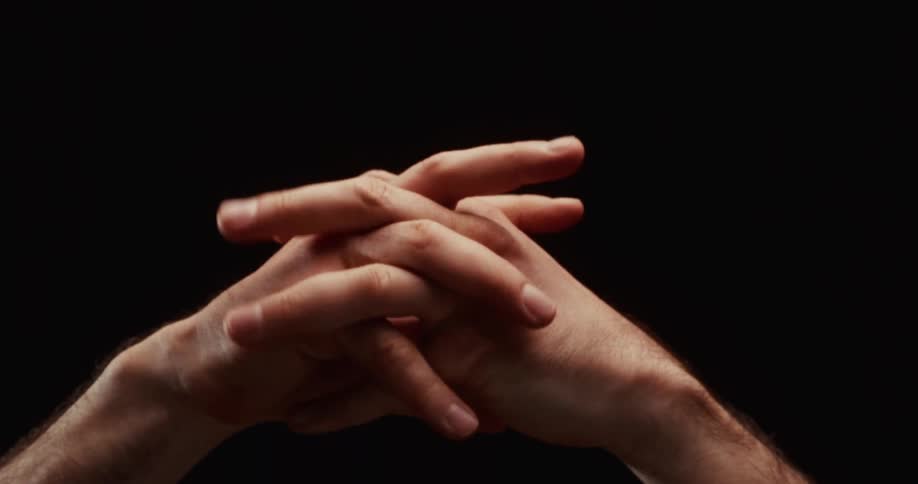 A dramatic slow motion close up of a man's hands slowly unclenching and clasping together against a black background