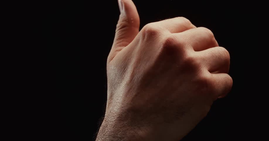 A close up of a man's hand in dramatic lighting slowly closing into a fist and then into a shaka hand gesture against a black background