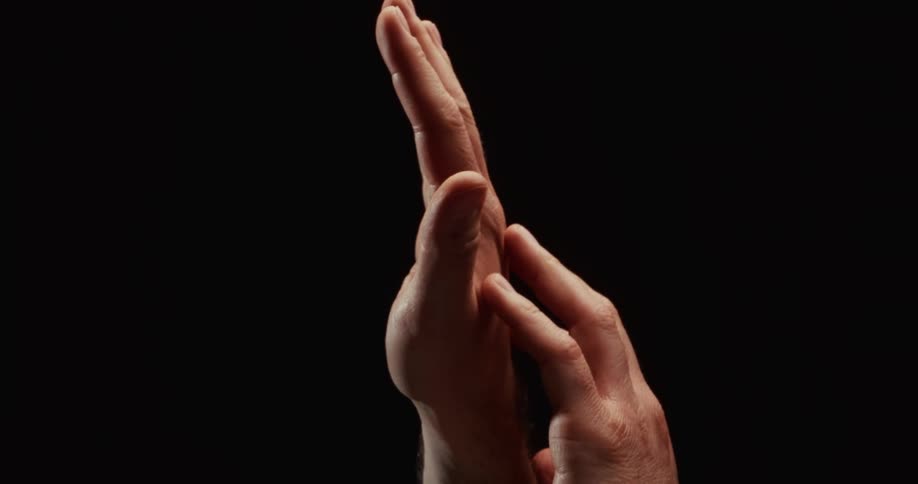 A man's hands slowly and gently caress each other in a dramatic, intimate close-up against a black background