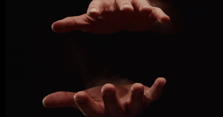 A man's hands slowly open and close in the darkness, with fine particles of dust or sand falling between them