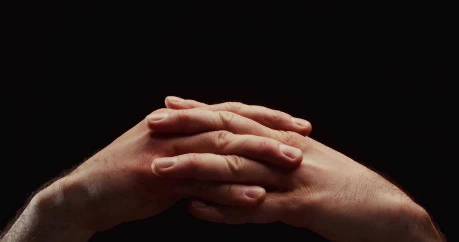 A dramatic close-up of hands clasped together in deep thought or prayer, slowly relaxing against a stark black background before separating