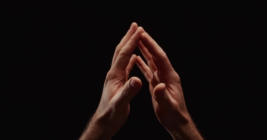 Dramatic slow motion close up of a man's hands clasped together in prayer against a solid black background