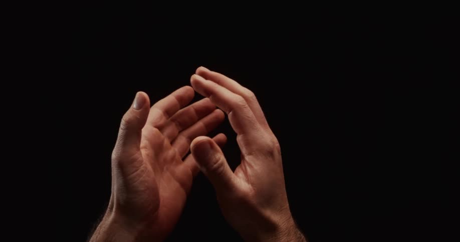 Male hands slowly come together in a prayer position against a dramatic black background
